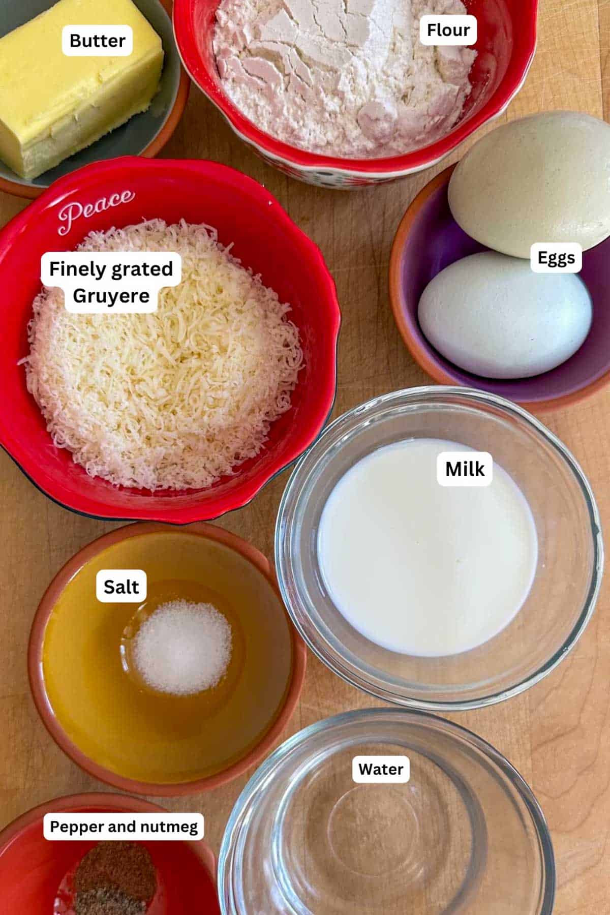 Ingredients needed to make Homemade Gruyere Drop Biscuits displayed in bowls including butter, flour, eggs, finely grated Gruyere, milk, salt, pepper and nutmeg, and water.