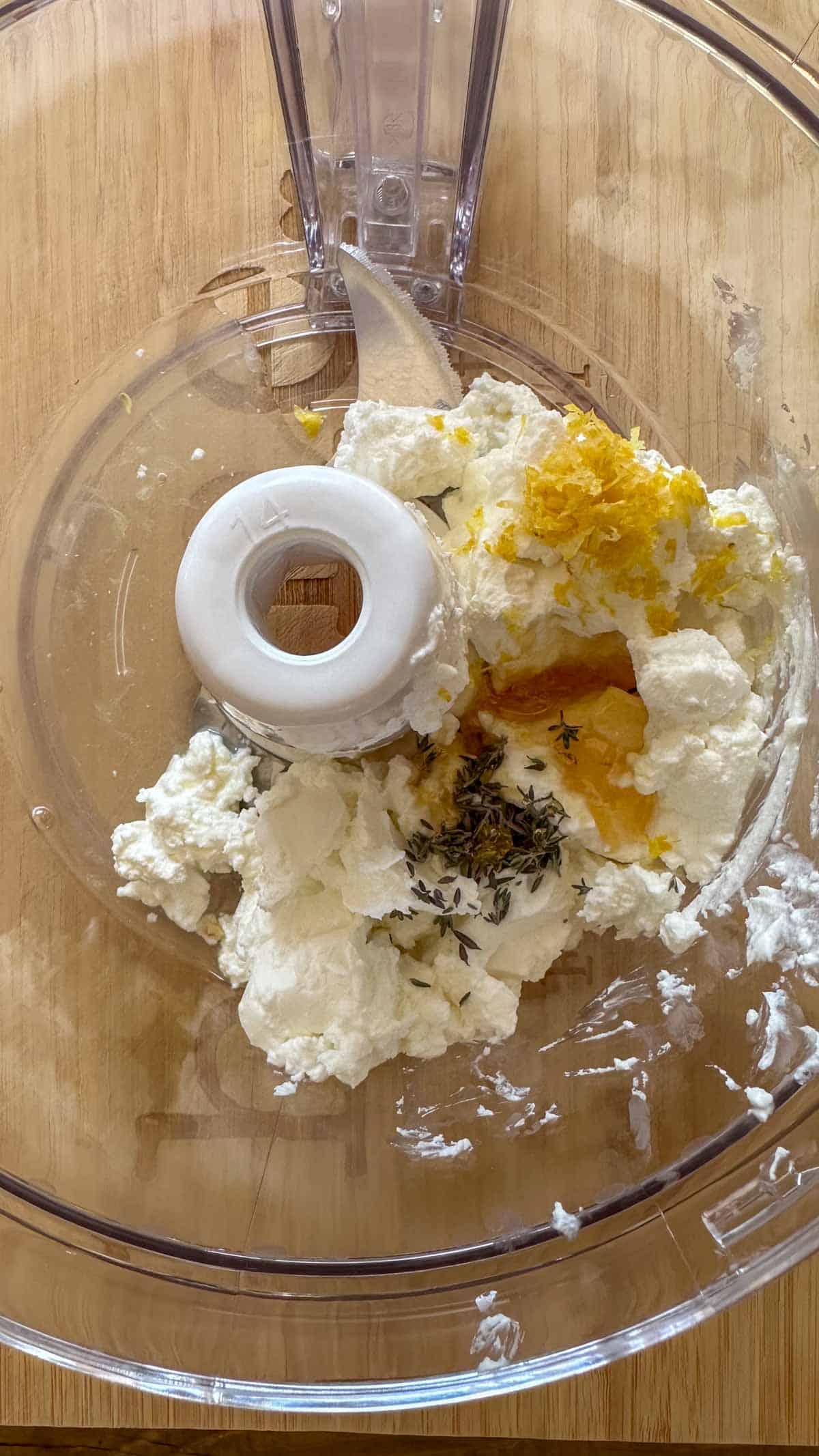 The bowl of a food processor containing the ingredients for Whipped Goat Cheese With Lemon Thyme Honey.