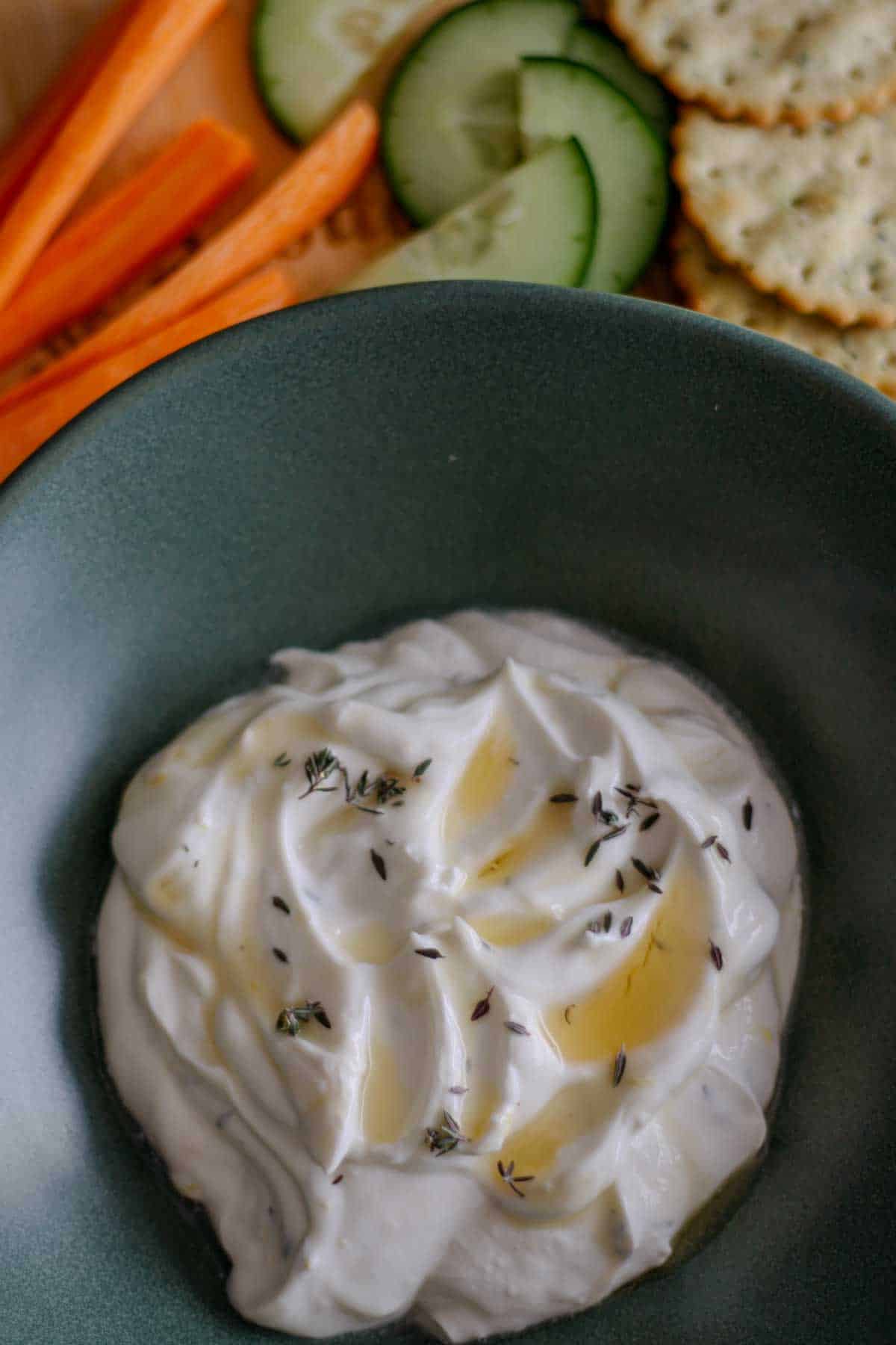 Whipped Goat Cheese With Lemon Thyme Honey in a green bowl, with carrot sticks, cucumber slices, and crackers in the background.