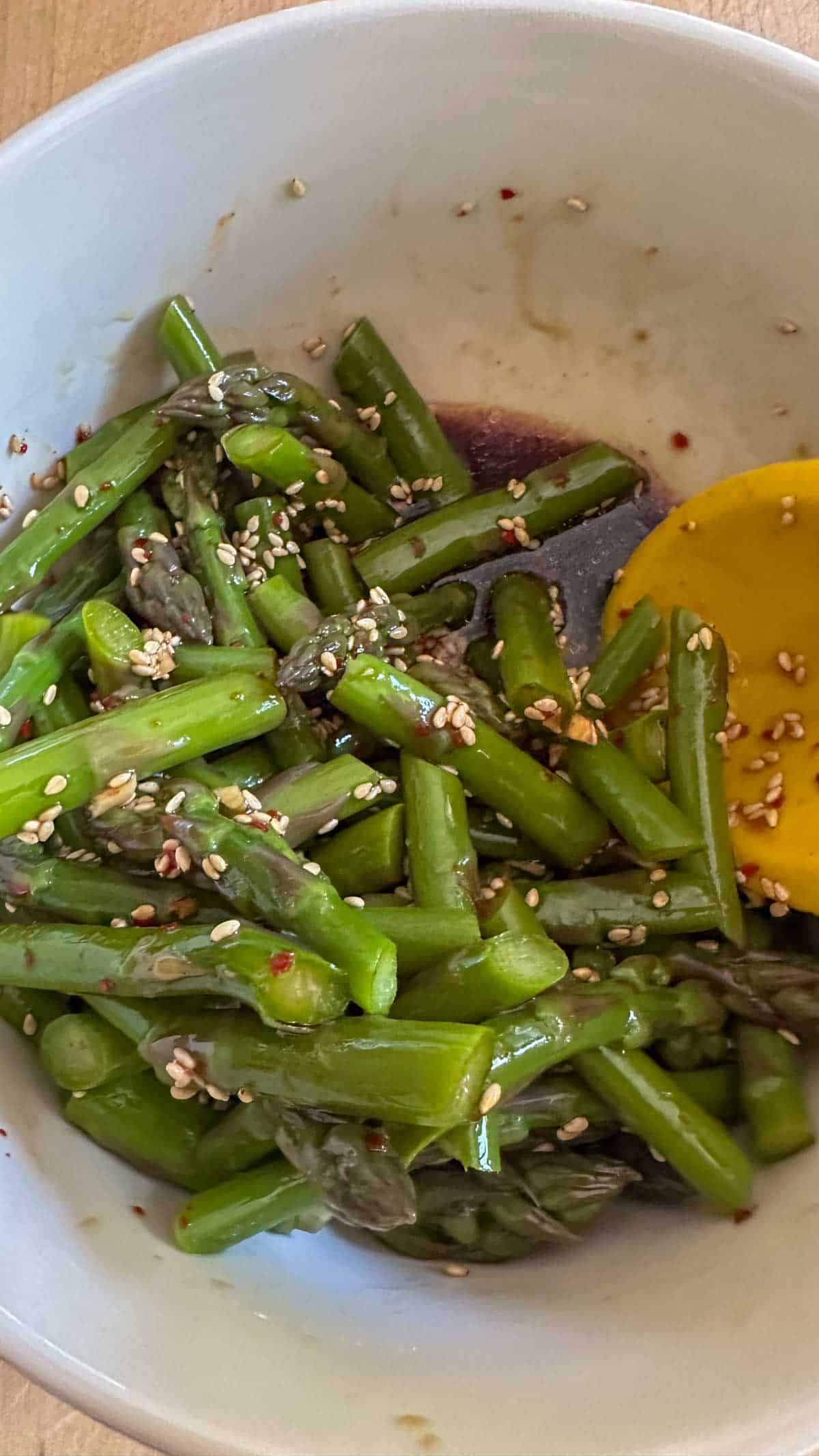 A bowl of asparagus with a soy sauce dressing, sesame seeds, and a spoon.