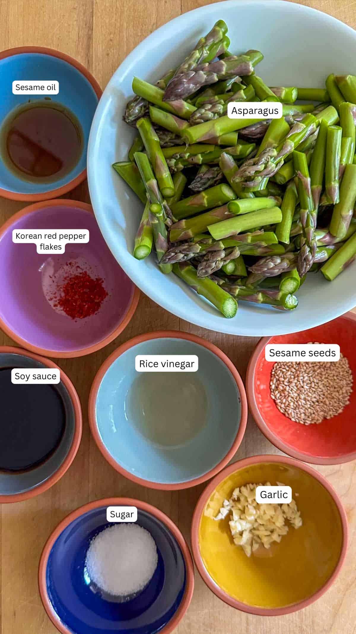 Ingredients for Korean cold asparagus in bowls including asparagus, sugar, sesame oil, Korean red pepper flakes, soy sauce, rice vinegar, sesame seeds, and garlic on a table.