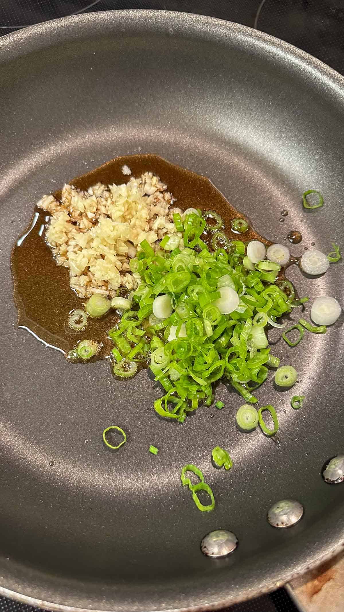 A nonstick skillet containing minced garlic, chopped green onions and sesame oil.