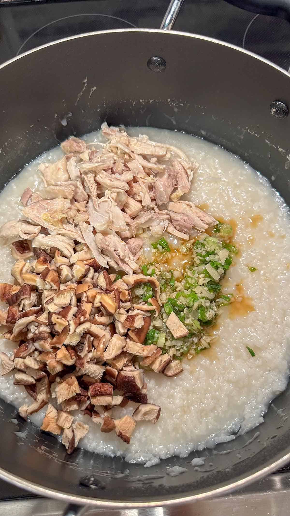 A large pot containing rice porridge with shredded chicken, chopped shiitake mushrooms, garlic, green onions, and sesame oil.