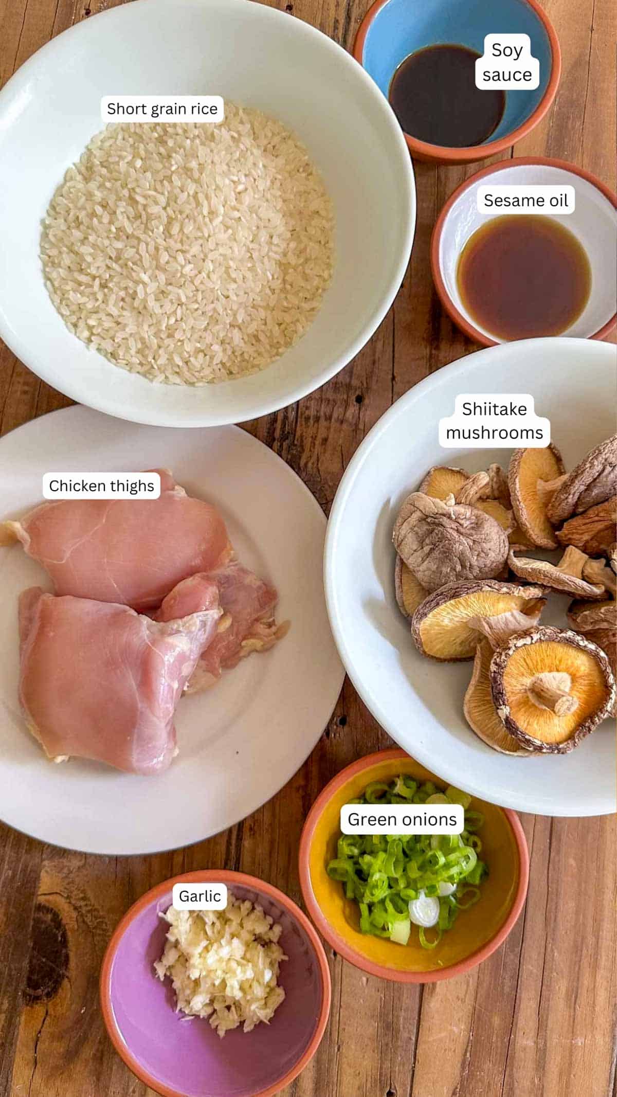 Ingredients for Juk With Chicken and Mushrooms including short grain rice, soy sauce, sesame oil, shiitake mushrooms, chicken thighs, green onions, and garlic displayed in bowls.