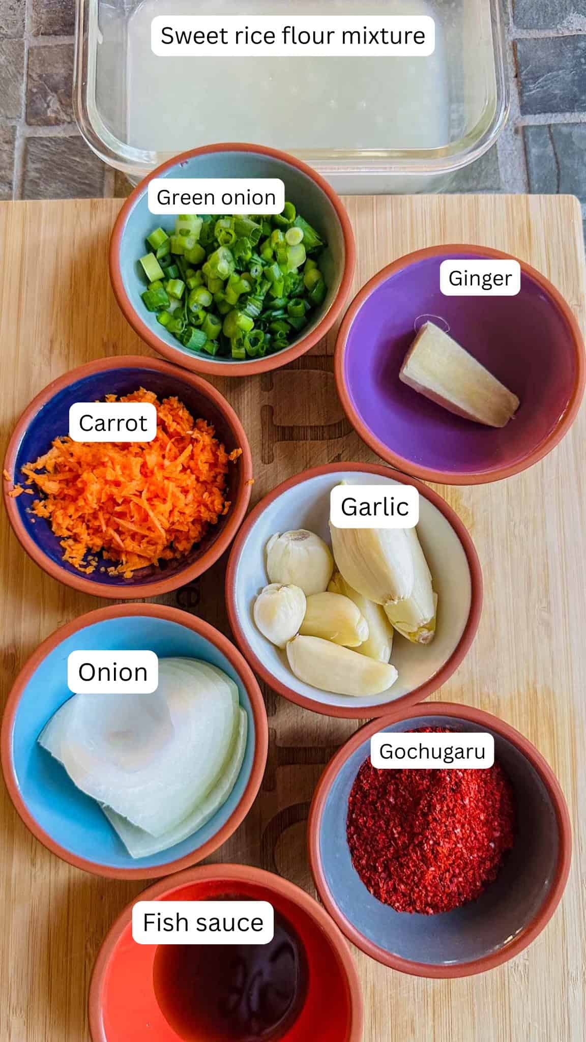 Ingredients to make kimchi including sweet rice flour mixture, and green onions, ginger, carrot, garlic, onion, gochugaru, and fish sauce displayed in bowls.