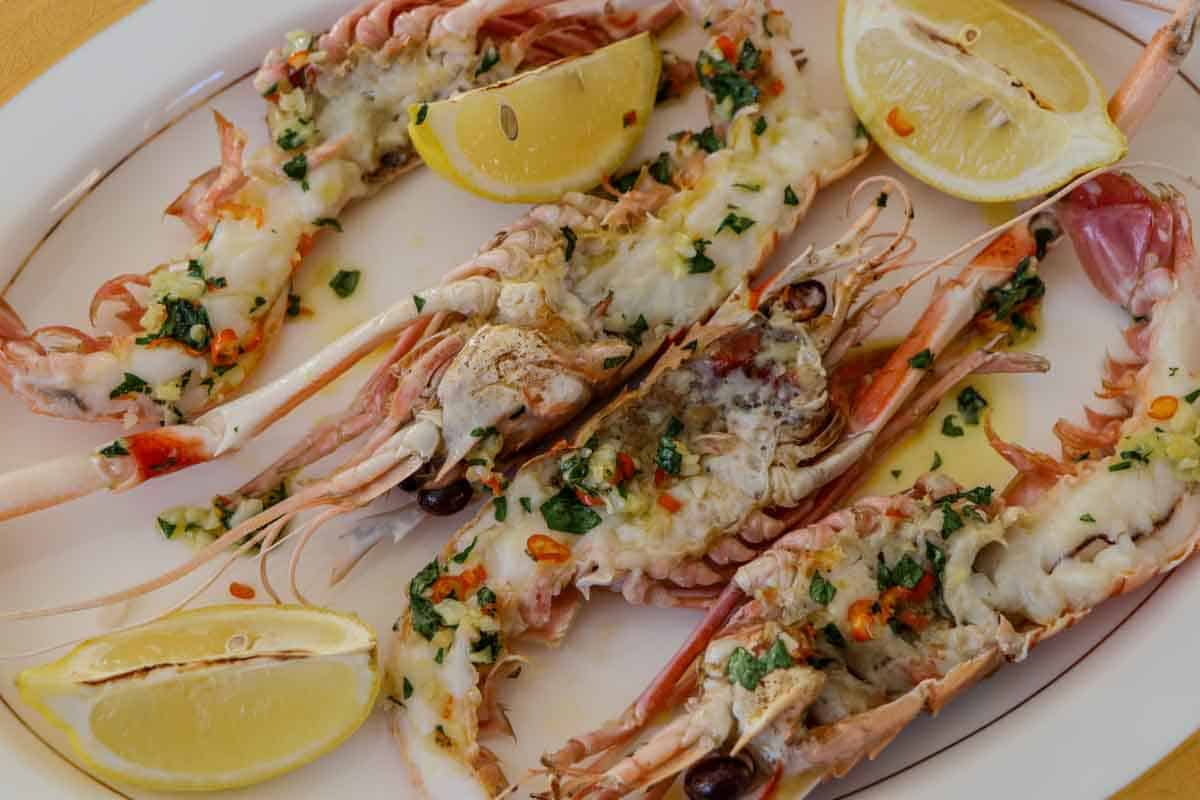 Broiled New Zealand scampi with garlic, chili, butter and parsley, served with lemon wedges on a gold rimmed plate.