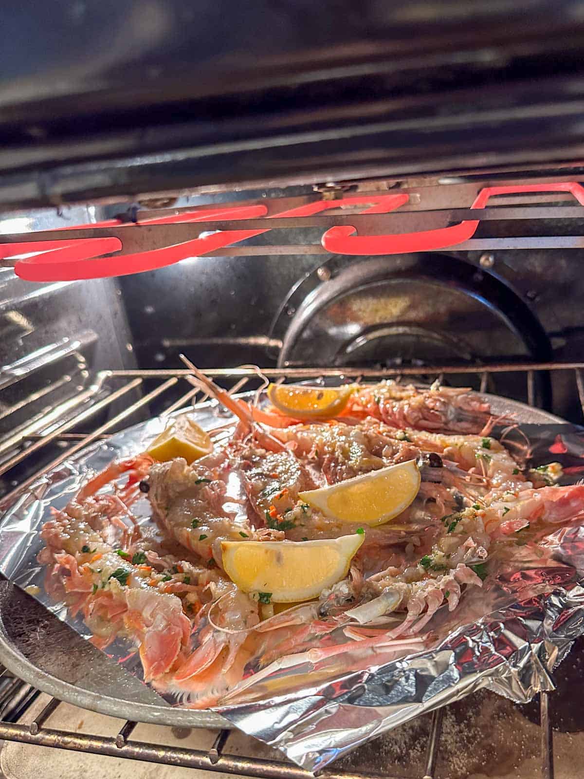 A baking tray lined with aluminum foil containing New Zealand scampi and lemon wedges under a broiler.