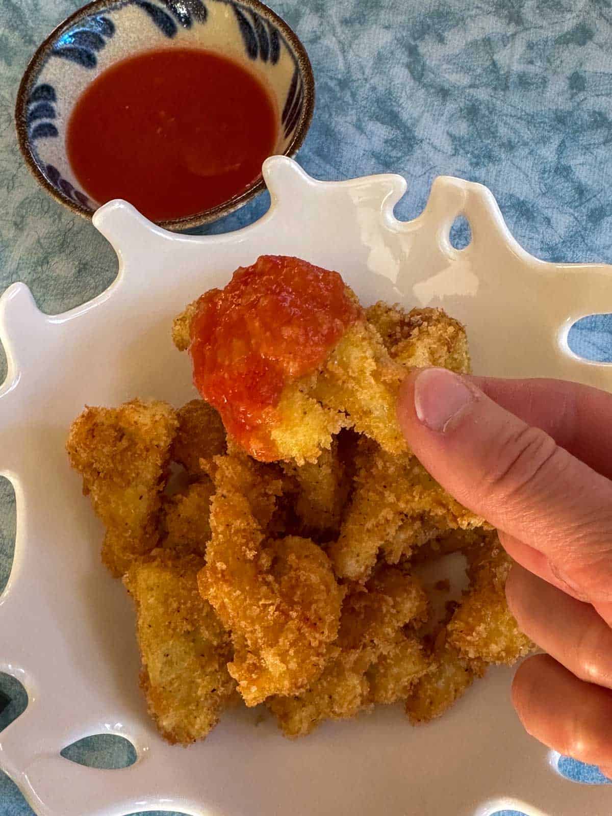 A white bowl containing fried cauliflower florets with a person holding one of the florets which was dipped in marinara sauce in the foreground, with a bowl containing the marinara in the background.