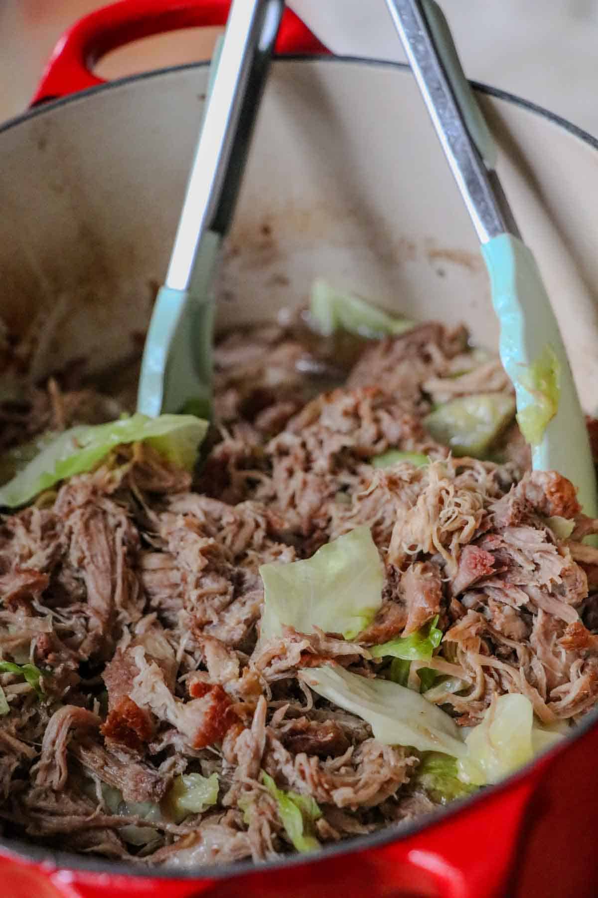 Tender shredded Kalua Pork with cabbage in a Dutch oven with a pair of tongs.