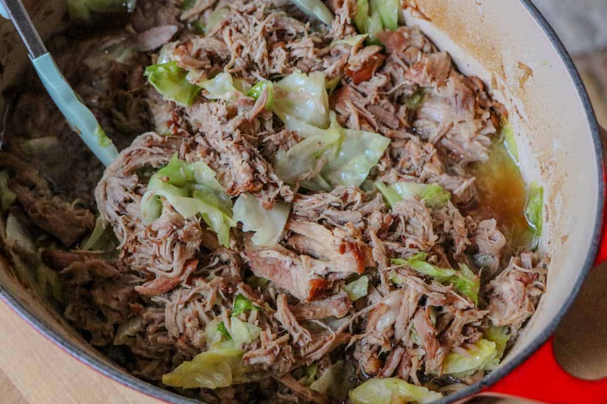 Tender shredded Kalua Pork with cabbage in a Dutch oven with a pair of tongs.
