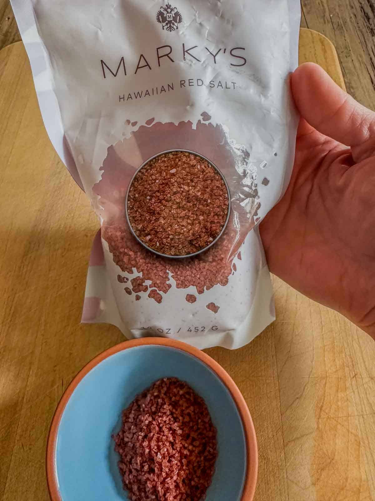 A small bowl with Hawaiian red salt and a bag of Marky's Hawaiian Red Salt held by a person's hand.