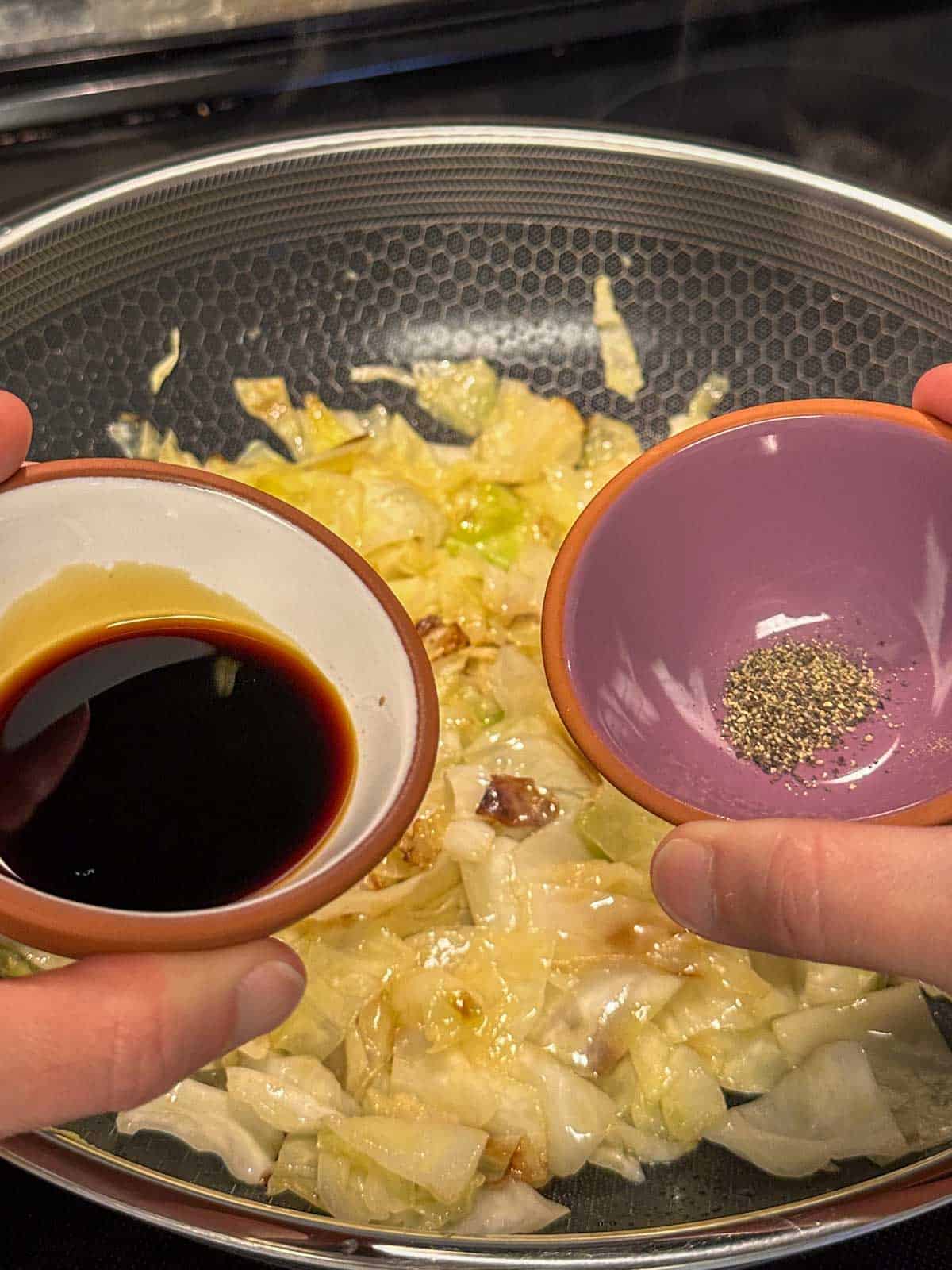 A wok containing sautéed cabbage in the background, and a person holding small bowls containing soy sauce and pepper in the foreground.