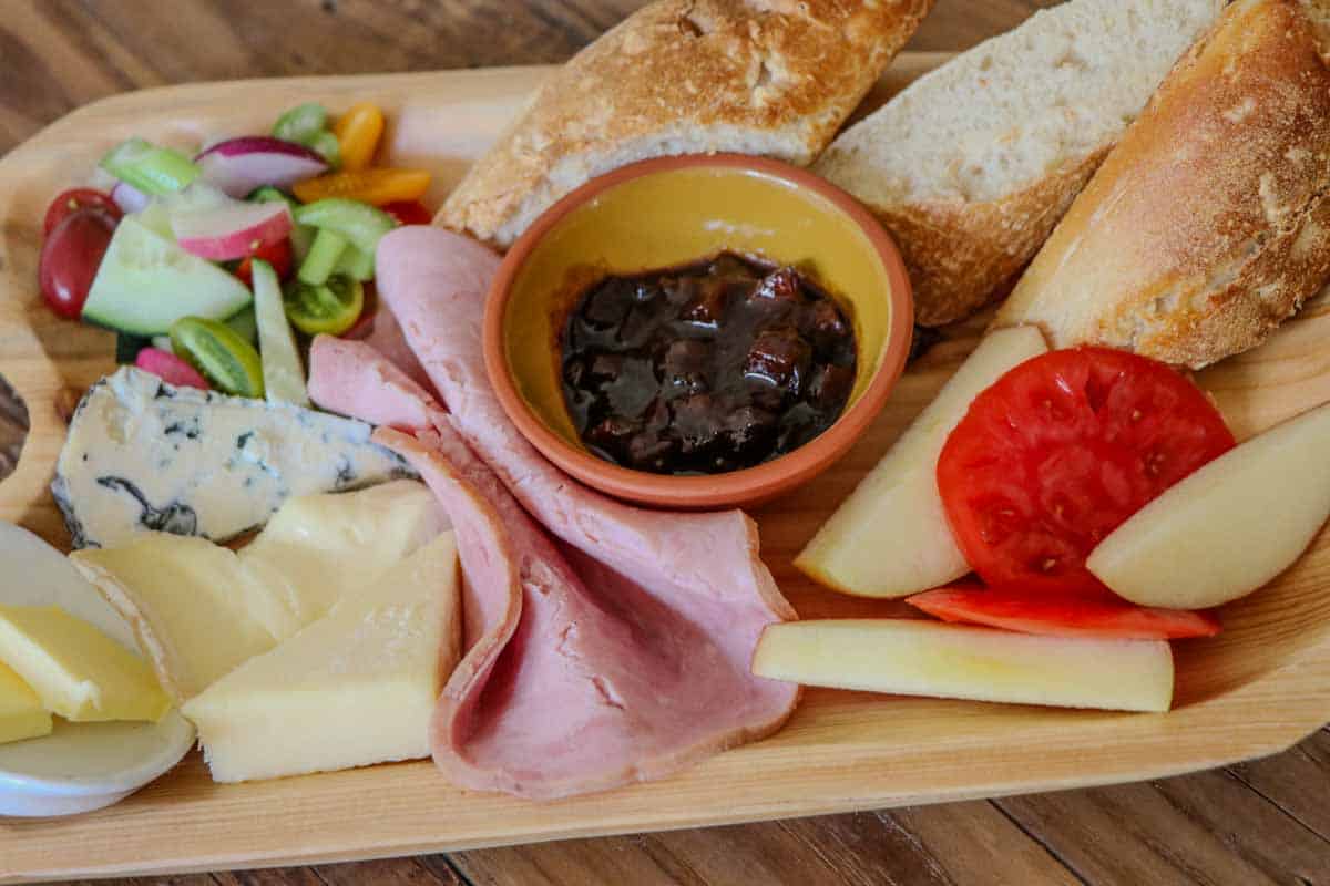 Ploughman's lunch displayed on a wooden platter including cheeses, ham, butter, vegetables, pieces of bread, Branston pickle in a bowl, sliced tomato and apple.