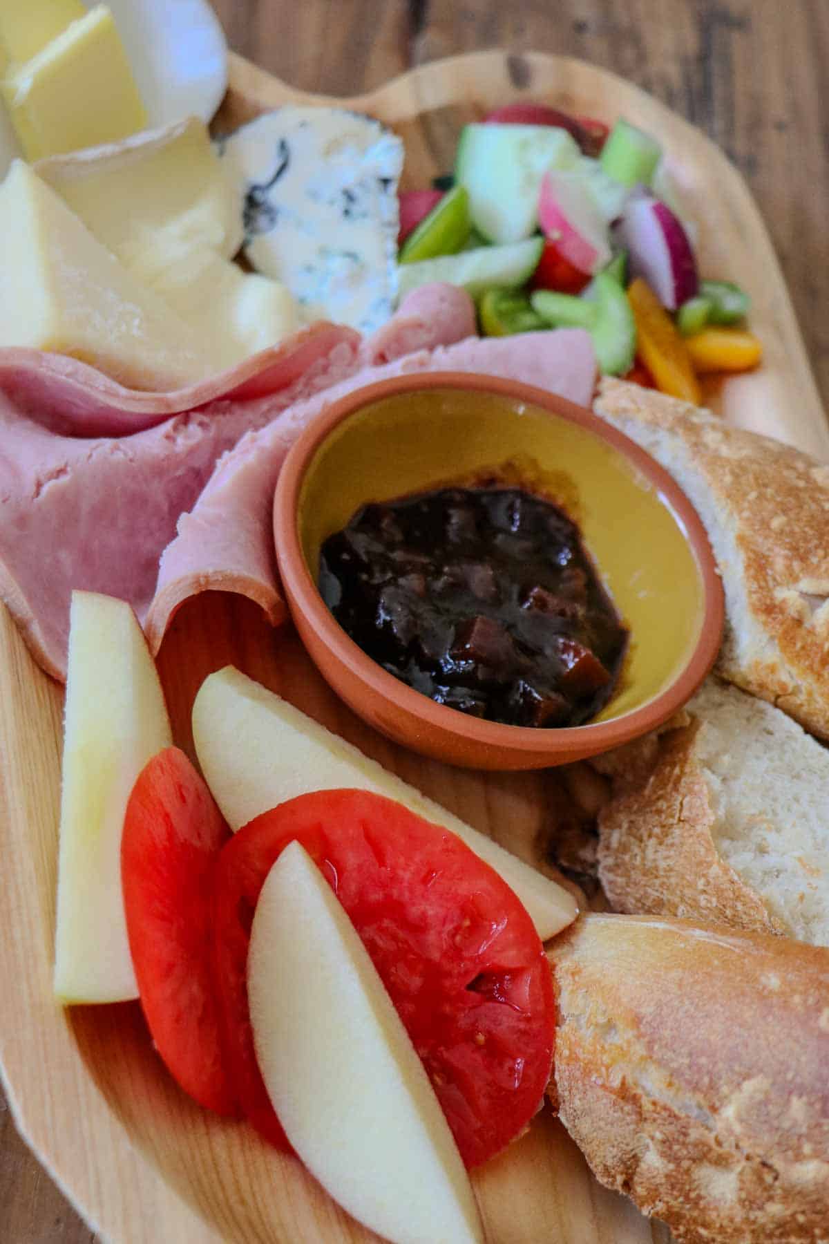 Ploughman's lunch displayed on a wooden platter including cheeses, ham, butter, vegetables, pieces of bread, Branston pickle in a bowl, sliced tomato and apple.
