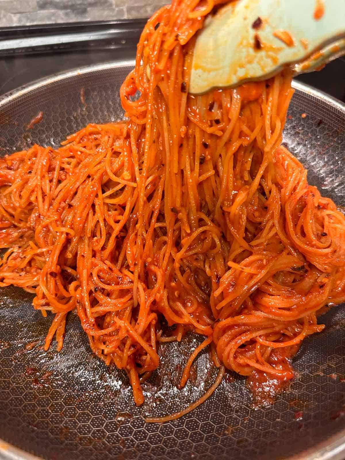 A skillet containing Spicy Angel Hair Pasta With Tomato Puree with a pair of tongs holding some of the pasta.