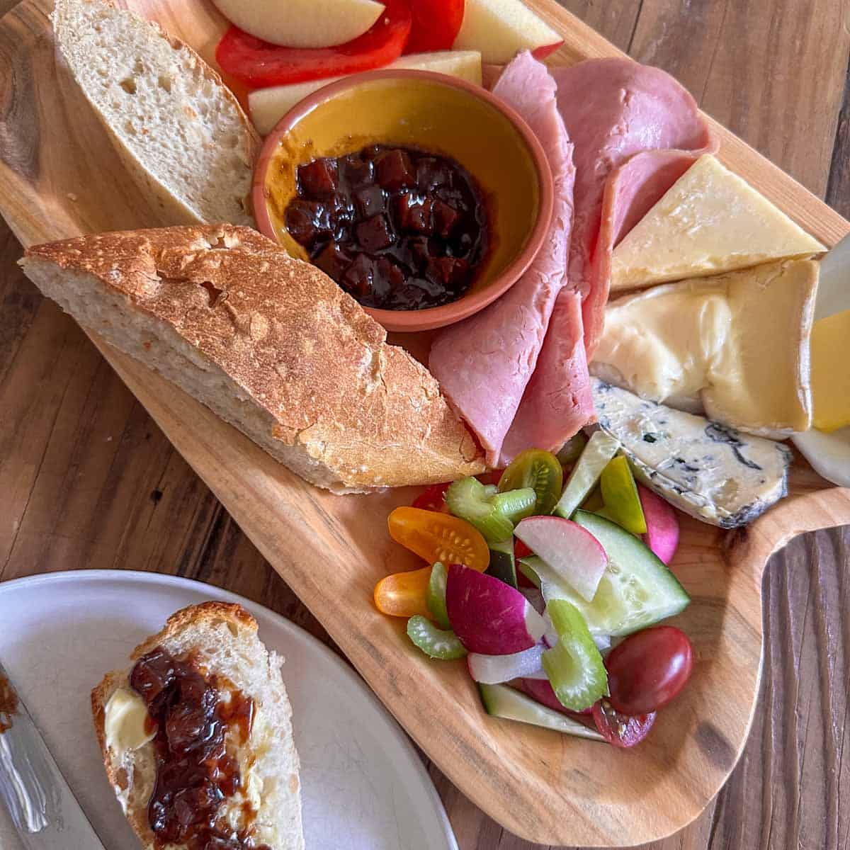 Ploughman's lunch displayed on a wooden platter including cheeses, ham, butter, vegetables, pieces of bread, Branston pickle in a bowl, sliced tomato and apple, next to a plate with a piece of bread slathered with butter and Branston pickled and a knife.