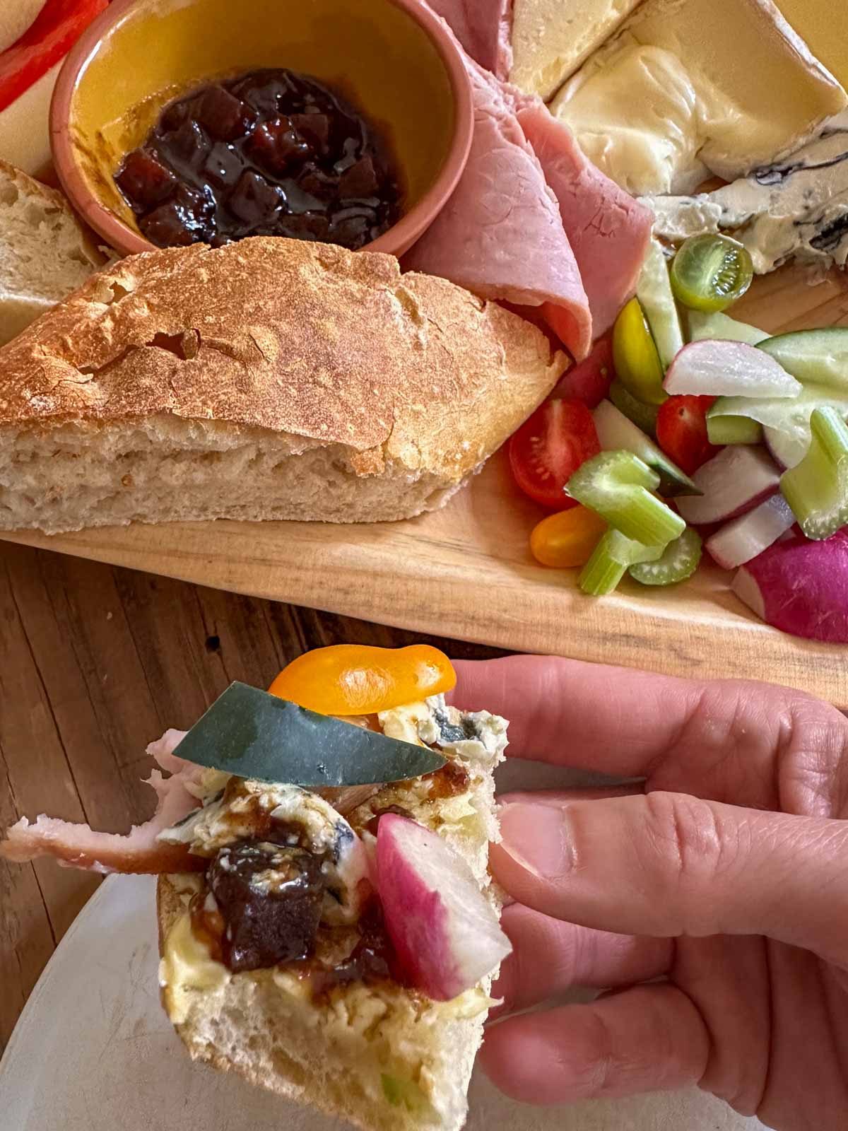 Ploughman's lunch displayed on a wooden platter including cheeses, ham, butter, vegetables, pieces of bread, Branston pickle in a bowl, sliced tomato and apple, and a person holding a piece of bread topped with butter, Branston pickle, ham, cheese, and vegetables.