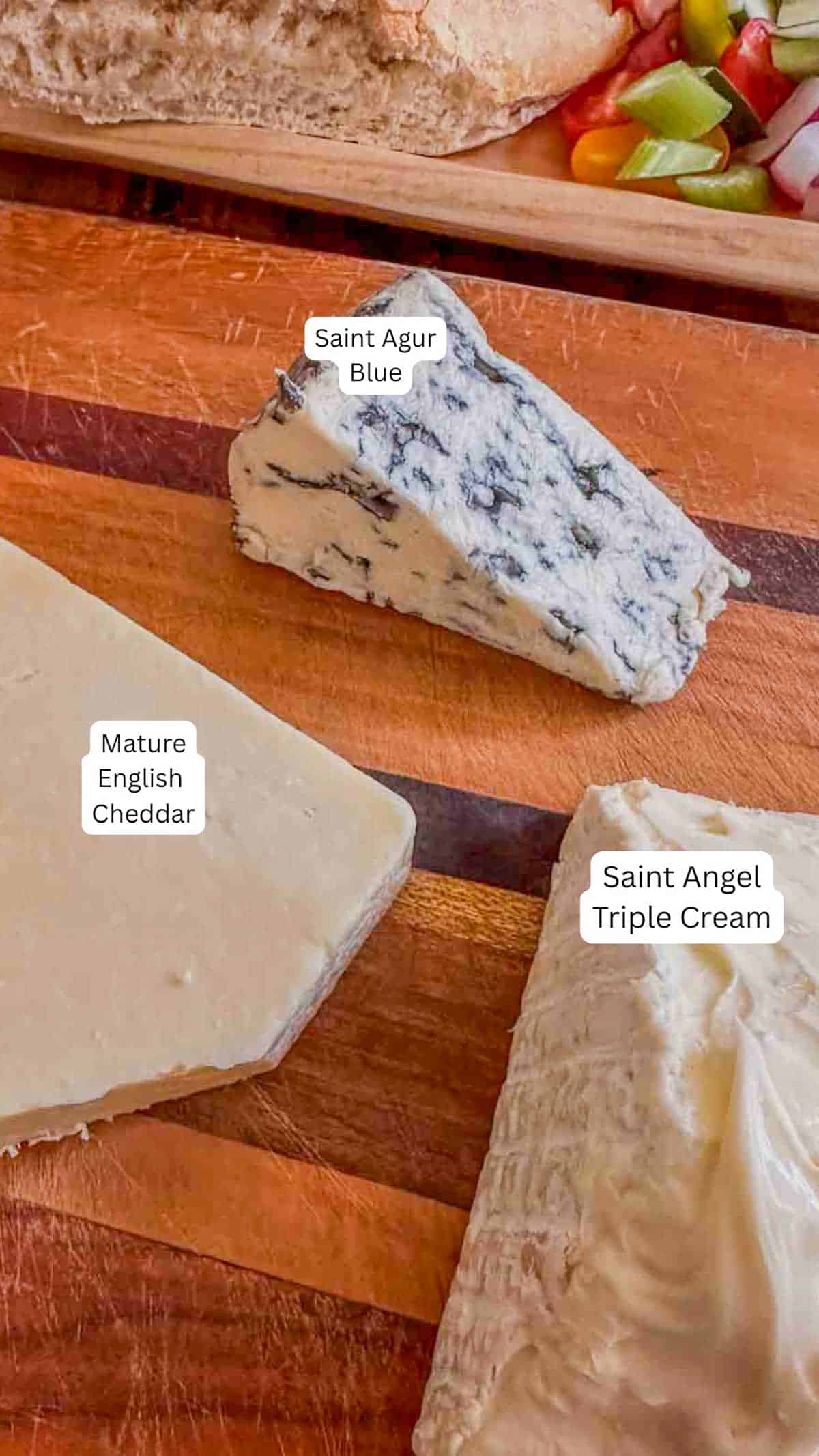 Cheddar, blue cheese, and triple cream cheese on a wooden cutting board, and bread and vegetables in the background.