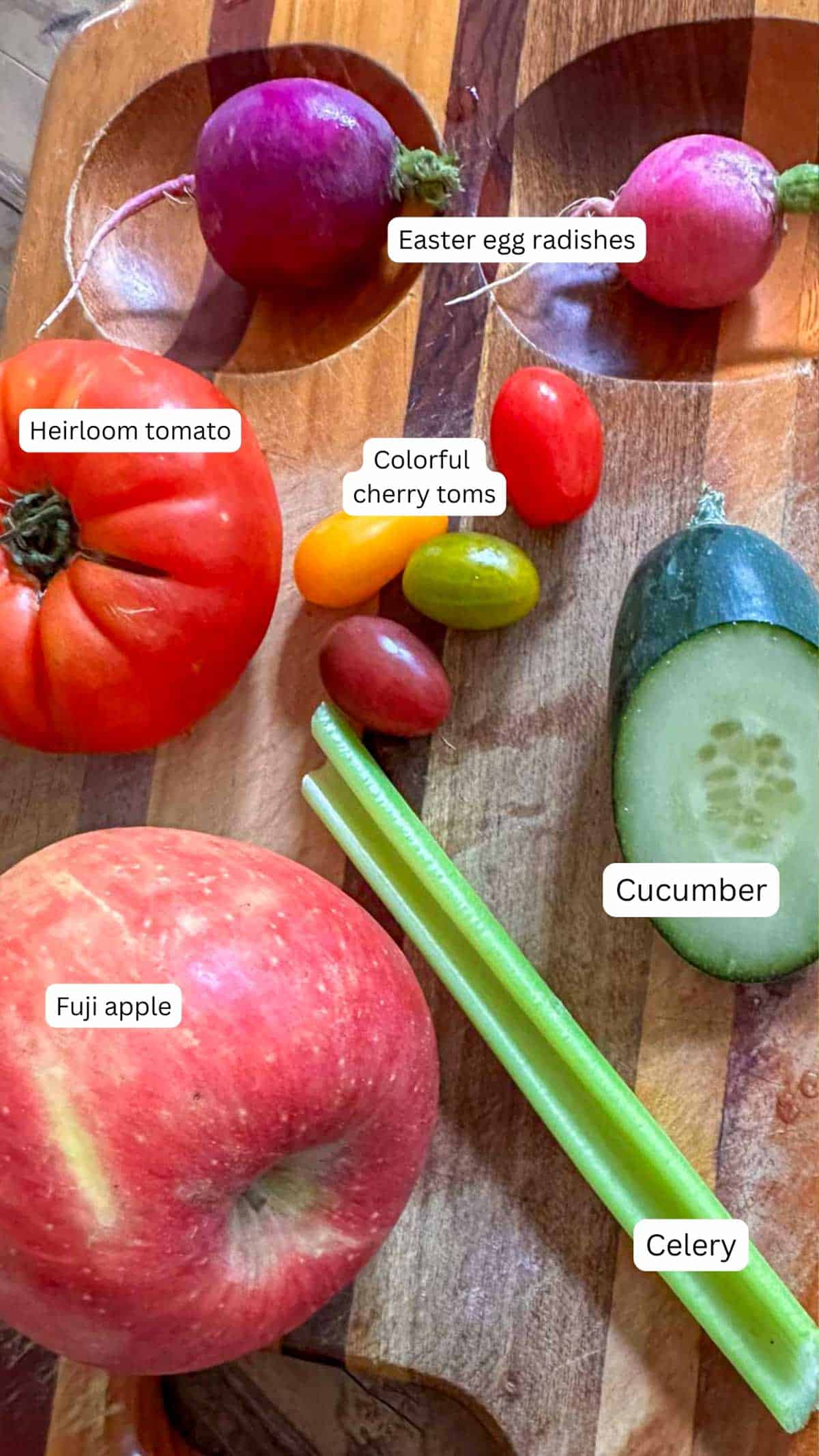 A wooden cutting board displaying an assortment of vegetables including a tomato, cherry tomatoes, celery, cucumber, radishes, and an apple.