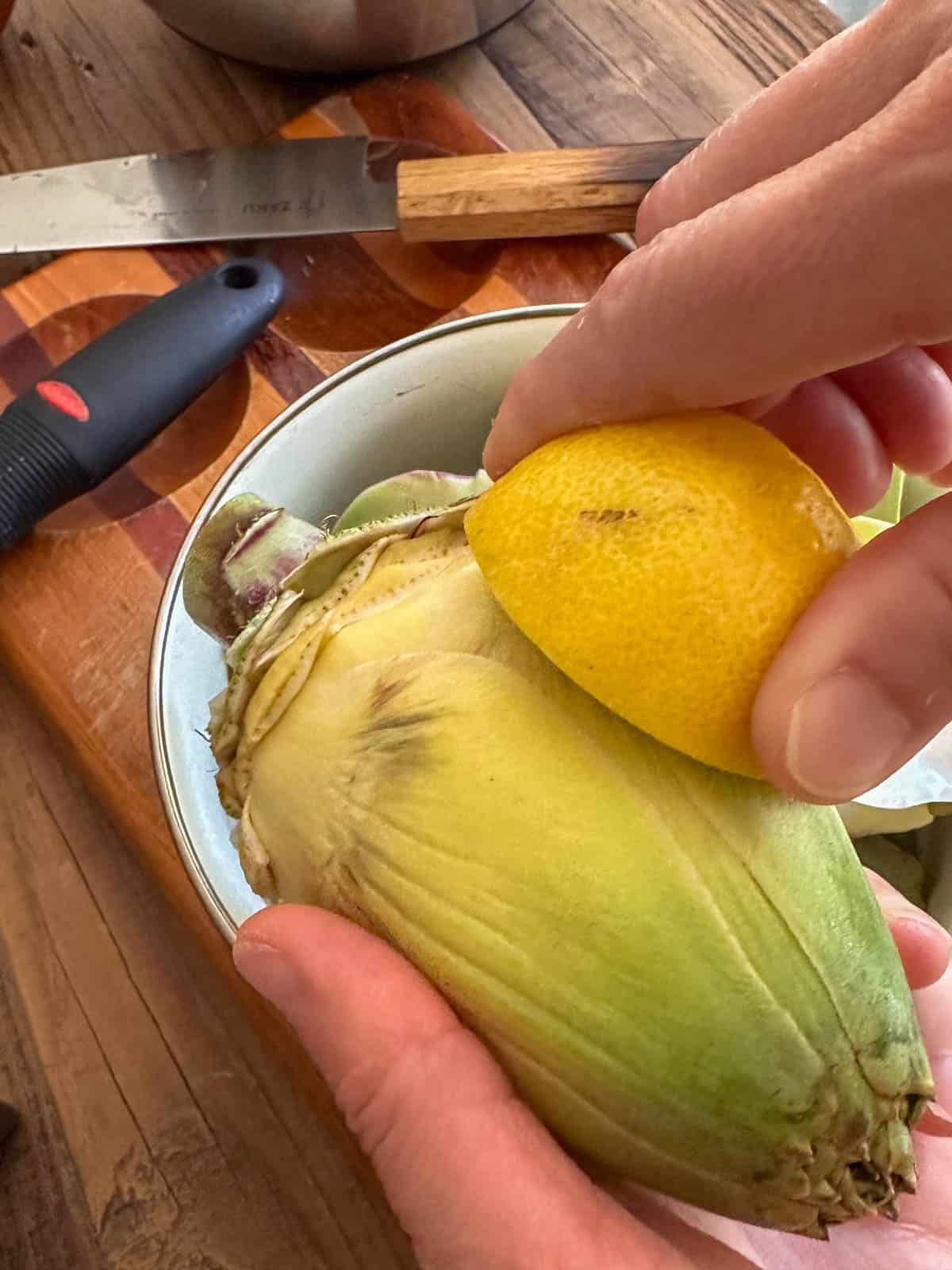 A person rubbing an artichoke with a lemon and a knife and peeler in the background.