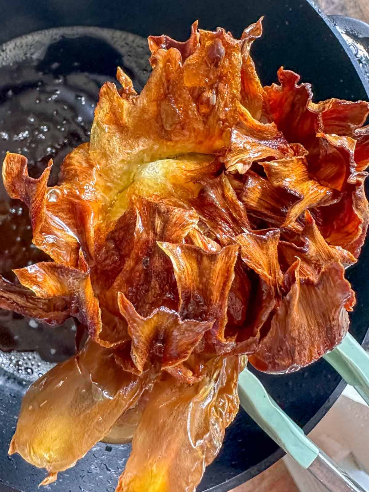 A pair of tongs holding a fried artichoke over a deep fat fryer filled with oil.