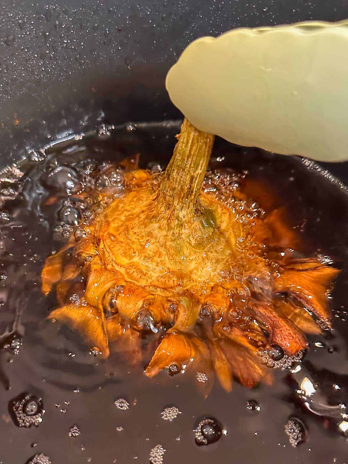 A pair of tongs holding an artichoke upside down frying in hot oil in a deep fryer.