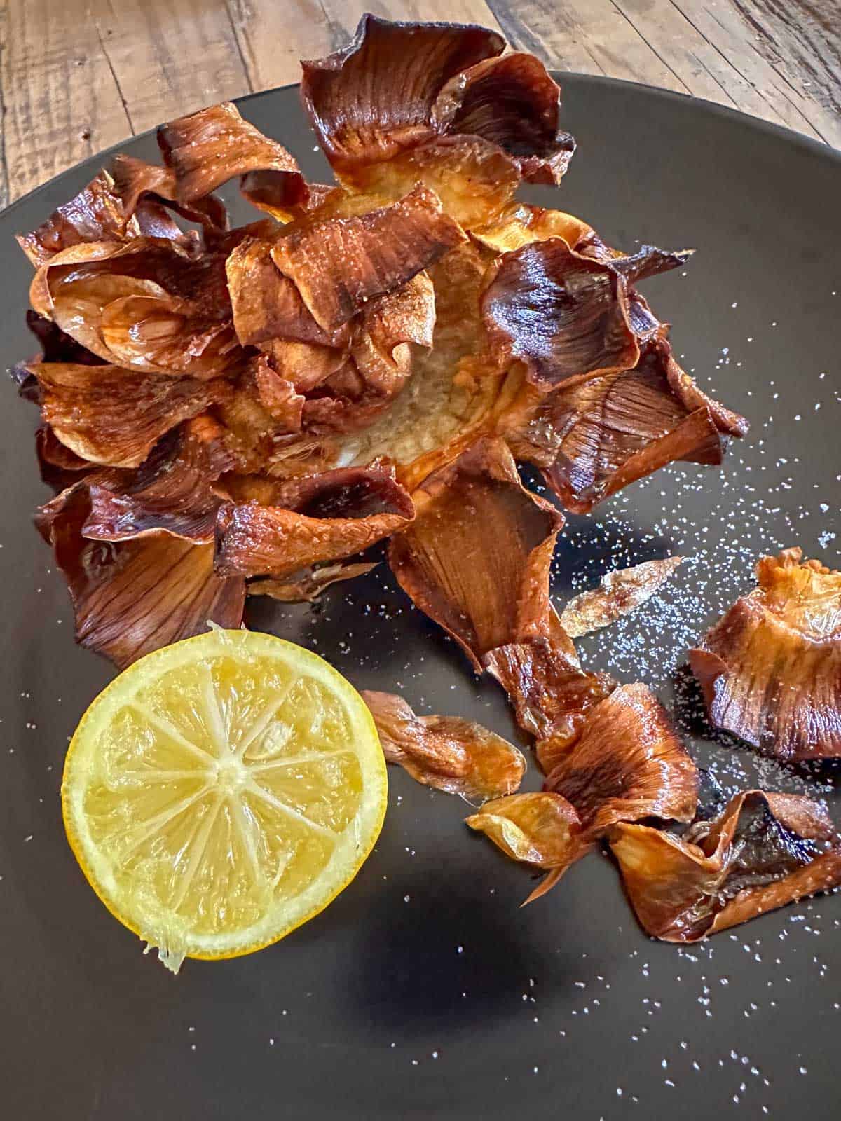 A black plate containing a crispy fried artichoke that has been salted and half a lemon.