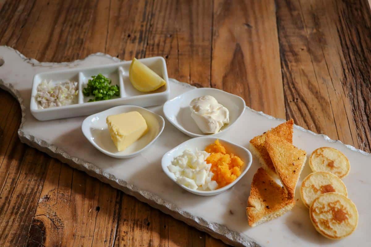 a serving plate with caviar toppings including lemon wedge, green onions, shallots, creme fraiche, chopped egg, toast points and blinis.