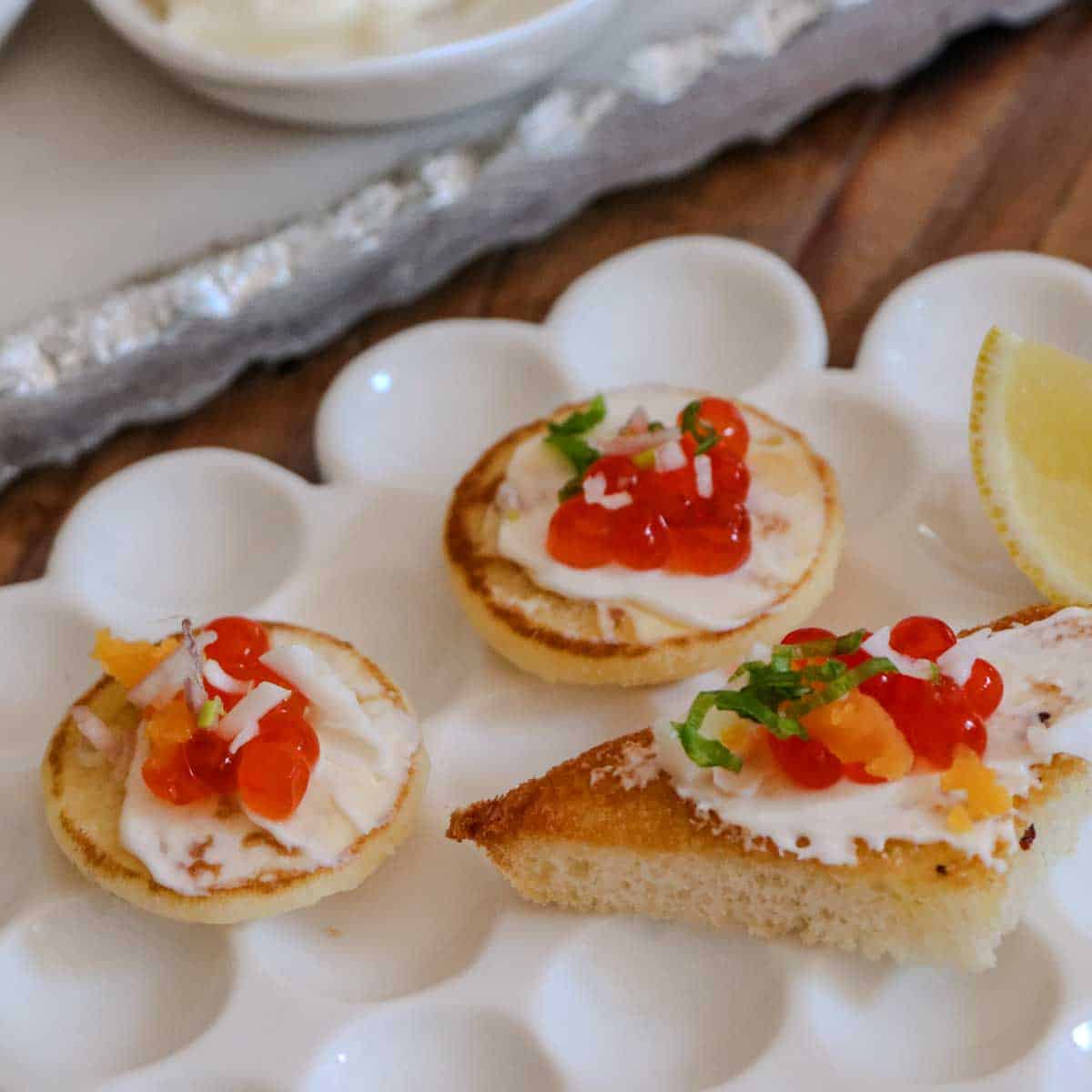A white plate topped with caviar appetizers including blinis and a toast point topped with creme fraiche, caviar, green onions, and shallots and a lemon wedge, with a part of another serving platter in the background.
