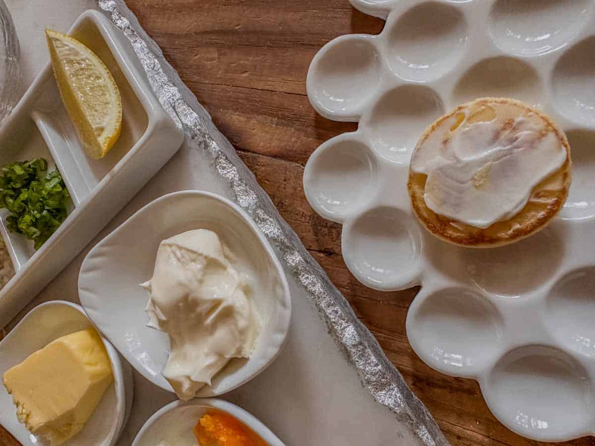 A white plate with dimples topped with a blini with creme fraiche, next to a serving tray with butter, creme fraiche, lemon, green onions, and chopped hard boiled egg.