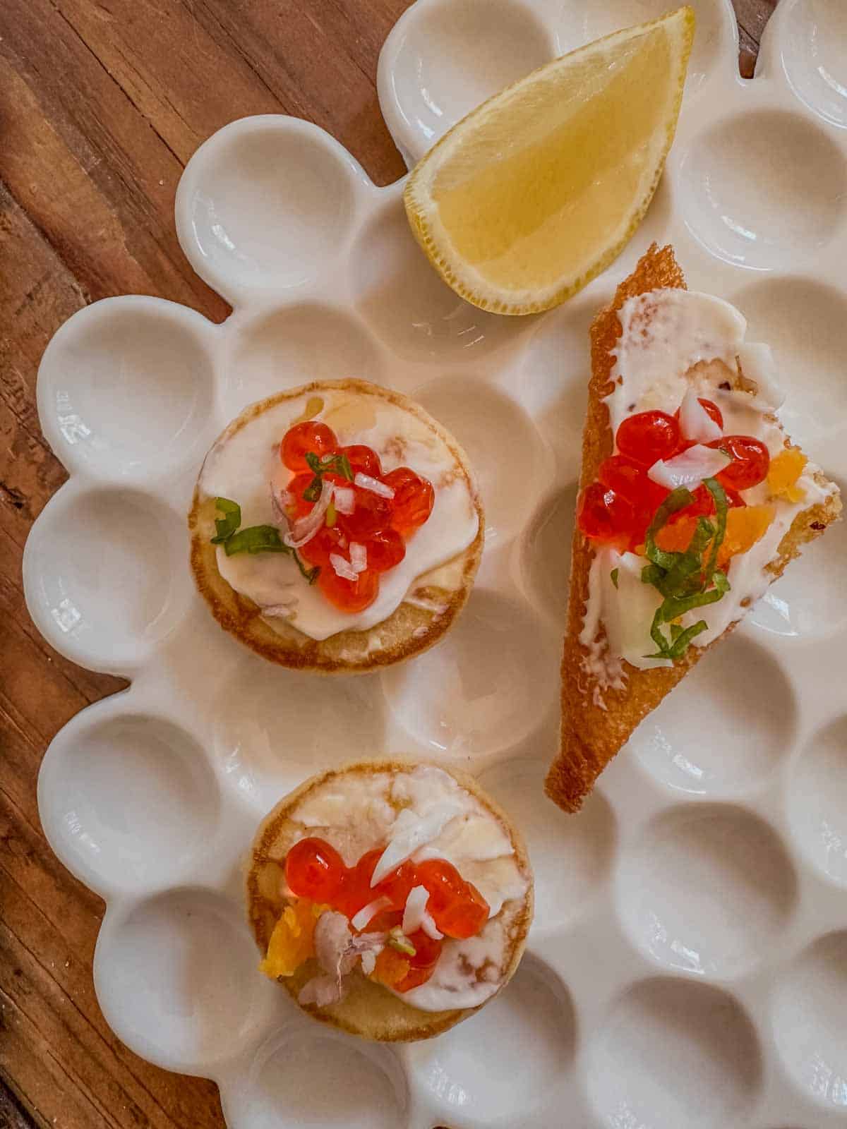 A white plate topped with caviar appetizers including blinis and a toast point topped with creme fraiche, cavair, green onions, and shallots and a lemon wedge.
