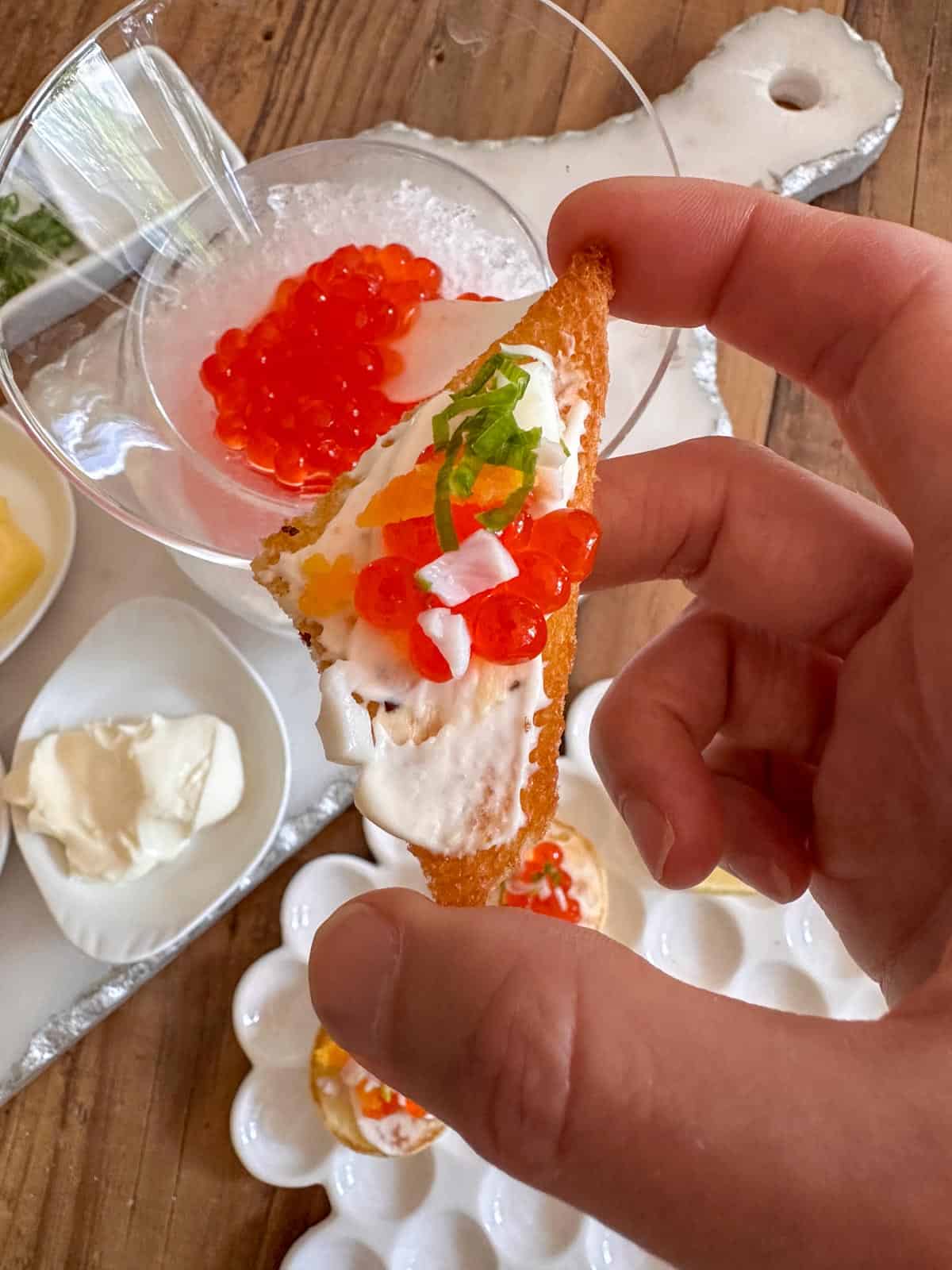 A hand holding a toast point caviar appetizer over a serving tray with a caviar filled caviar serving dish and a bowl of creme fraiche, and another plate with more caviar appetizers.